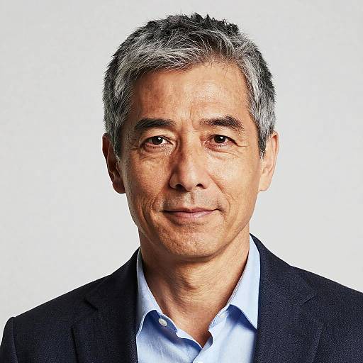 Photograph of middle-aged Asian man with short gray hair, wearing a black blazer and light blue shirt, smiling against a white background.