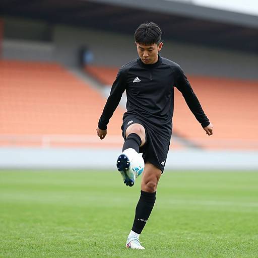 Photograph of an Asian male soccer player in black long-sleeve kit and black shorts, mid-kick, on a green field with orange stadium