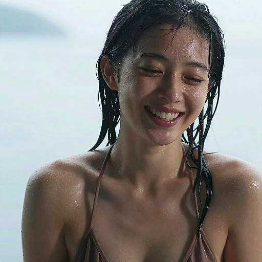 Photograph of a smiling Asian woman with wet, dark hair, wearing a brown bikini top, against a bright, cloudy sky background.