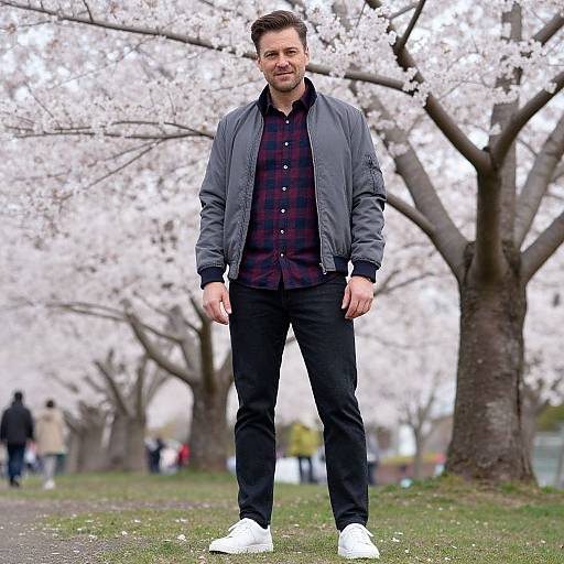 Photograph of a bearded man with short brown hair, wearing a gray jacket, red plaid shirt, black pants, and white sneakers, standing