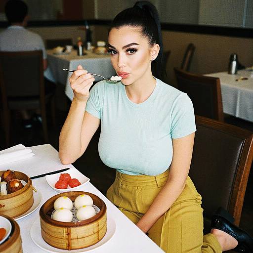 Photograph of a dark-haired woman with fair skin, wearing a light blue top and mustard-yellow pants, eating from a steamed bun at a dim