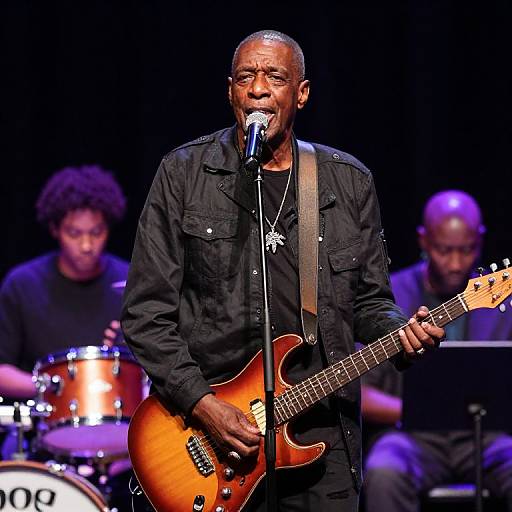 Photograph of an older African-American man with short hair, wearing a black shirt, singing into a microphone while playing a sunburst electric guitar on stage