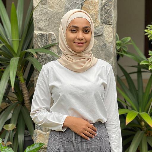 Photograph of a young woman with medium brown skin, wearing a beige hijab, white long-sleeve shirt, and gray plaid skirt,
