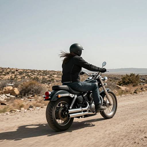 Photograph of a person in black leather jacket and helmet riding a black motorcycle on a dusty desert road under clear blue sky.