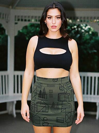 Photograph of a young woman with medium skin tone, dark hair, wearing a black crop top with a cut-out and a green patterned mini-sk
