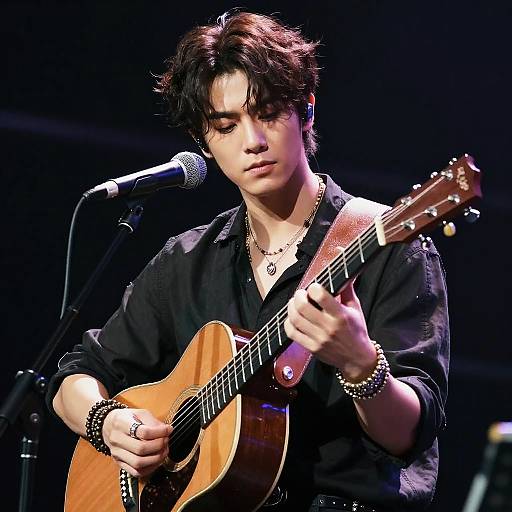 Photograph of Asian male musician with messy black hair, wearing black shirt and bracelets, playing acoustic guitar on stage.