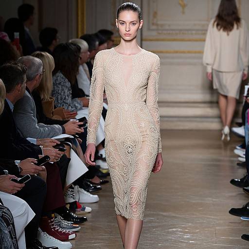 Photograph of a slim, dark-haired model in a sheer, gold lace long-sleeve dress, walking a fashion runway with seated audience.
