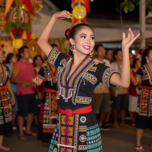 Photograph of an Asian woman dancing in a traditional, colorful, embroidered dress with red sash, black background, festive crowd, and vibrant decorations.