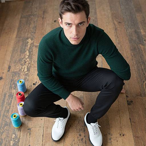 Photograph of a young man with short dark hair, green sweater, black pants, and white sneakers, squatting on wooden floor beside colorful yarn sp