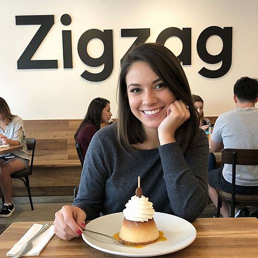 Photograph of a smiling young woman with dark hair, wearing a black sweater, enjoying a topped dessert in a brightly lit café with 