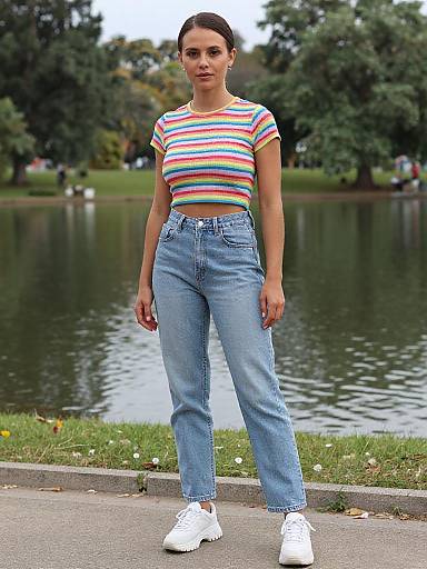 Photograph of a woman with dark hair in a striped rainbow top and high-waisted blue jeans, standing by a park lake. She wears white