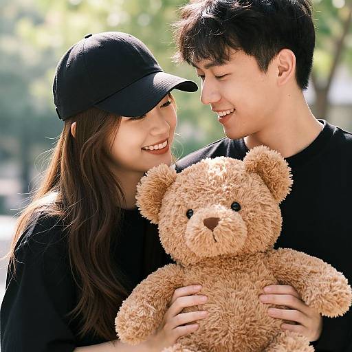 Photograph of a smiling Asian couple in black clothes, holding a fluffy brown teddy bear, outdoors with blurred greenery background.