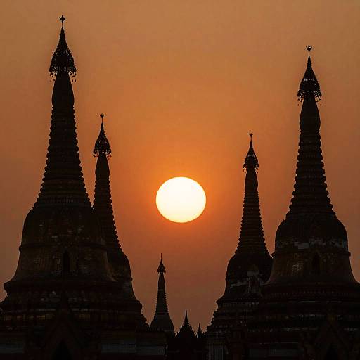 Photograph of a vibrant orange sunset behind the silhouetted peaks of ancient, ornate pagodas, creating a striking contrast between the glowing