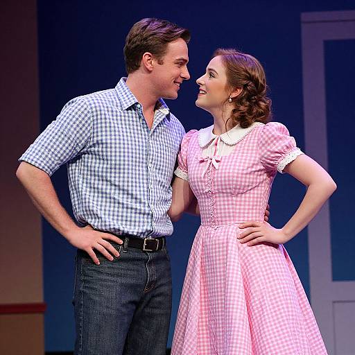 Photograph of a smiling couple on stage; man in blue check shirt, dark jeans, woman in pink check dress with white collar.