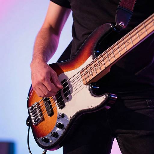 Photograph of a male musician's hands playing a sunburst electric bass guitar, wearing a black shirt, with a blurred blue background.
