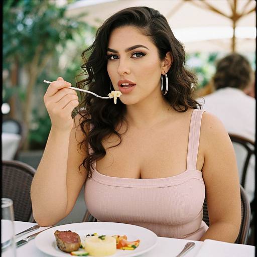 Photograph of a curvy, dark-haired woman with hoop earrings, wearing a pink tank top, eating a meal outdoors with a fork.