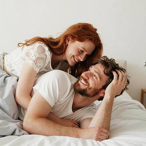 Photograph of a smiling red-haired woman in a white lace top, hugging a bearded man in a white t-shirt, both lying in bed
