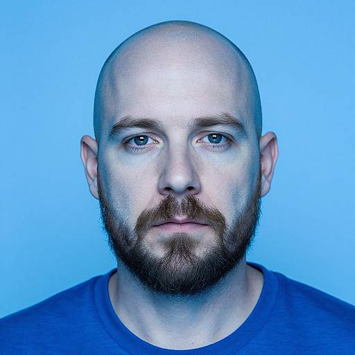 Photograph of a bald, bearded Caucasian man with blue eyes, wearing a blue shirt, against a bright blue background.