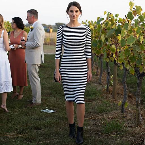 Photograph of a woman in a black-and-white striped dress and black boots standing in a vineyard, with three other people in the background.