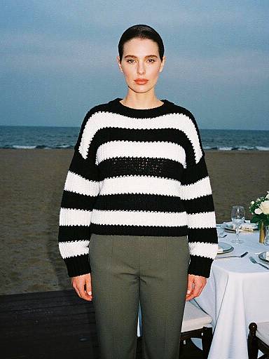 Photograph of a woman with dark hair in a black and white striped sweater, olive pants, standing at a beachside table.
