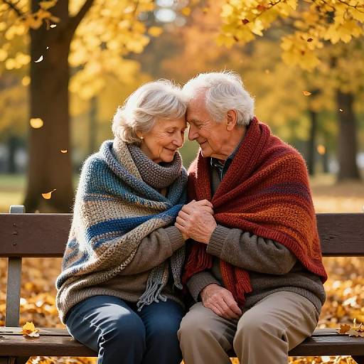 Photograph of an elderly couple sitting on a bench in an autumn park, smiling and holding hands, wrapped in colorful blankets.