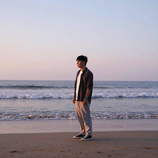 Photograph of a young Asian man standing on a beach at sunset, wearing a black jacket, white shirt, beige pants, and black sneakers, looking