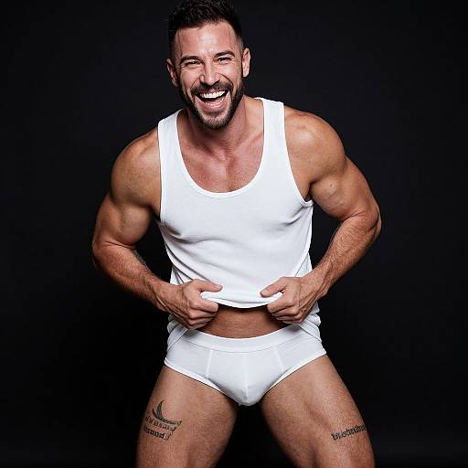 Photograph of a muscular, bearded man with short dark hair, laughing, wearing a white tank top and briefs, black background, tattoos on