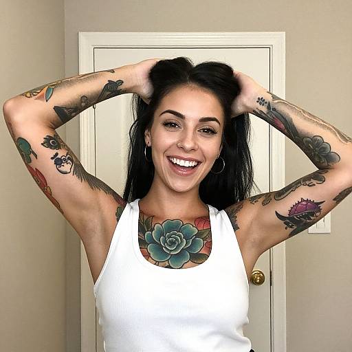 Photograph of a smiling woman with black hair, tattoos on arms and chest, wearing a white tank top, hands in hair, standing in front of