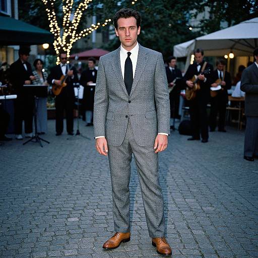 Photograph of a handsome man in a gray checkered suit, black tie, and brown leather shoes, standing on a cobblestone street at an