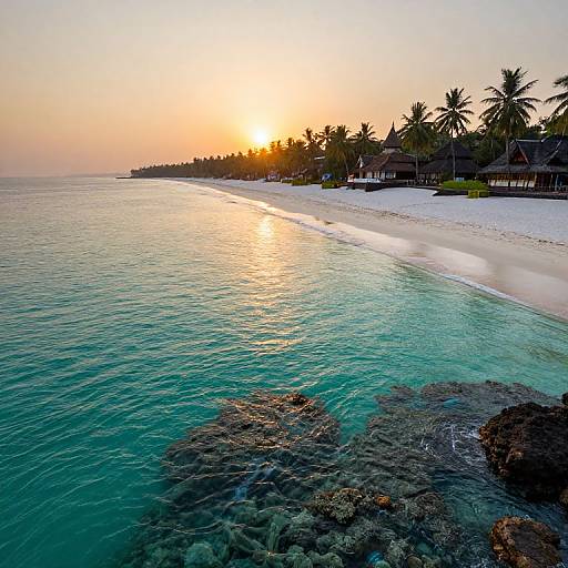 Photograph of a tropical beach at sunset with clear turquoise water, rocky shoreline, palm trees, and thatched huts along the white sand.