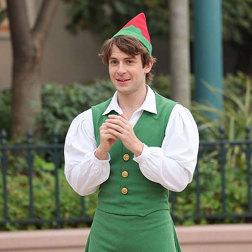 Photograph of a young white man with brown hair, wearing a green elf costume with a red-green hat, white shirt, and green vest, smiling