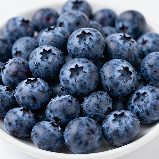 Close-up photograph of fresh, plump blueberries with dew drops on their dark blue, speckled skins, piled in a white bowl.