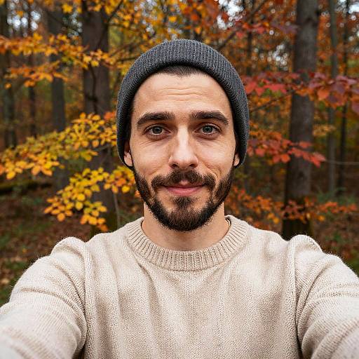 Photograph of a bearded man with dark hair, wearing a black beanie and cream sweater, standing in a forest with autumn-colored leaves in the