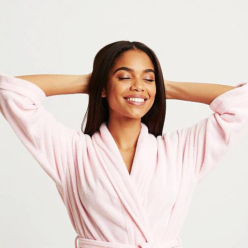 Photograph of a smiling young woman with medium brown skin and straight black hair, wearing a white bathrobe, with arms raised behind her head, against