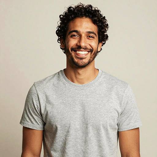 Photograph of a smiling, curly-haired man with a beard, wearing a light gray t-shirt, standing against a plain white background.