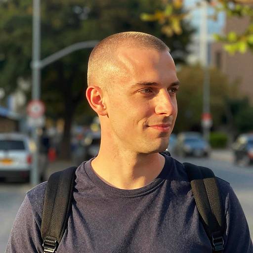 Photograph of a bald, light-skinned man with a short beard, wearing a dark blue shirt and black backpack, standing on a sunlit city