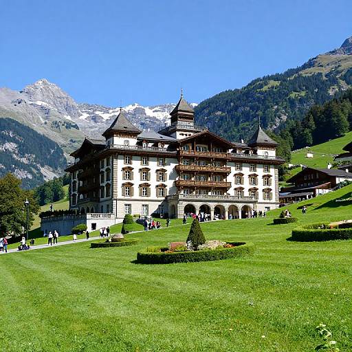 Photograph of a grand, white, multi-story mountain lodge with dark wooden balconies, set in a lush green meadow, surrounded by pine trees