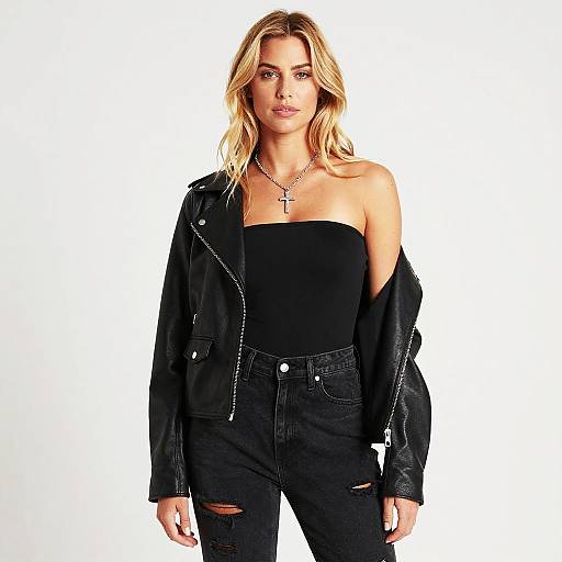 Photograph of a blonde woman with wavy hair, wearing a black strapless top, black ripped jeans, and a black leather jacket, standing against