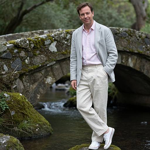 Photograph of a smiling man with short brown hair, wearing a light gray blazer, white shirt, white pants, and white sneakers, standing on
