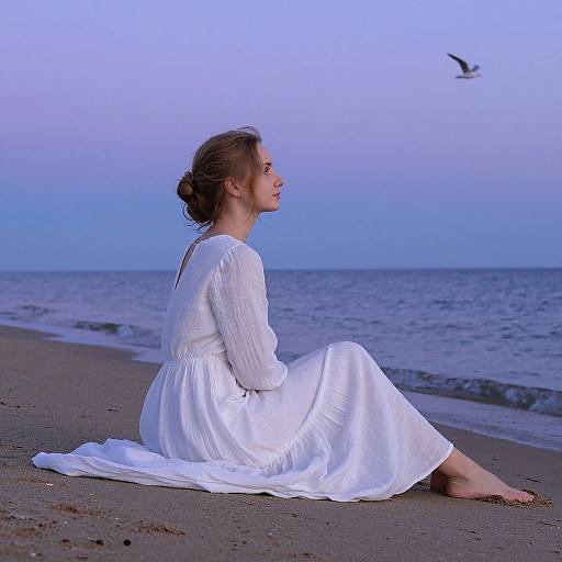 Photograph of a woman with brown hair in a bun, wearing a white, long-sleeved, flowing dress, sitting on a beach at twilight