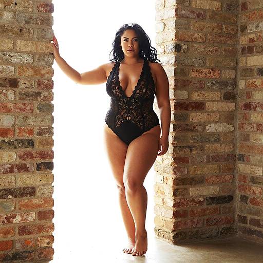 Photograph of a curvy, dark-haired woman in a black lace bodysuit, standing in a brick doorway, backlit, barefoot,