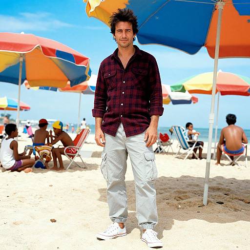 Photograph of a dark-haired man in a maroon plaid shirt and white cargo pants standing on a sunny beach with colorful umbrellas and people in