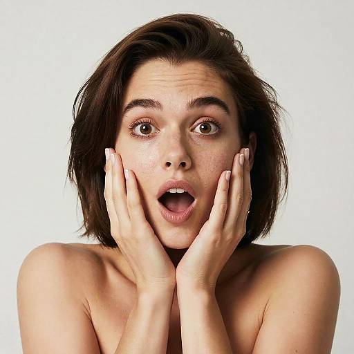 Photograph of a surprised young woman with short brown hair, wide brown eyes, and open mouth, hands covering her cheeks, against a white background.