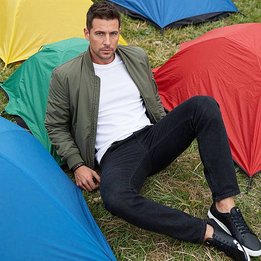 Photograph of a handsome, dark-haired man in a green jacket, white t-shirt, black jeans, and black sneakers, sitting among colorful camping tents