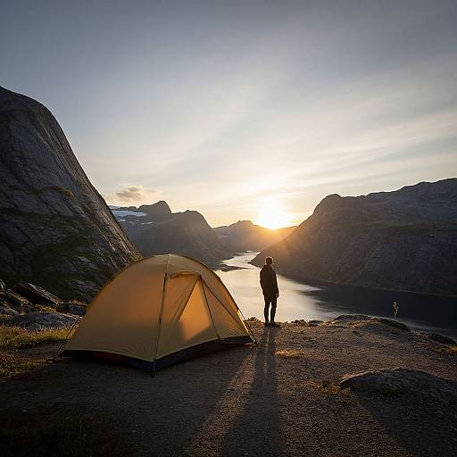 Photograph: Silhouetted camper stands beside yellow tent at sunset, overlooking serene lake surrounded by rocky mountains, with sunlight reflecting on water.