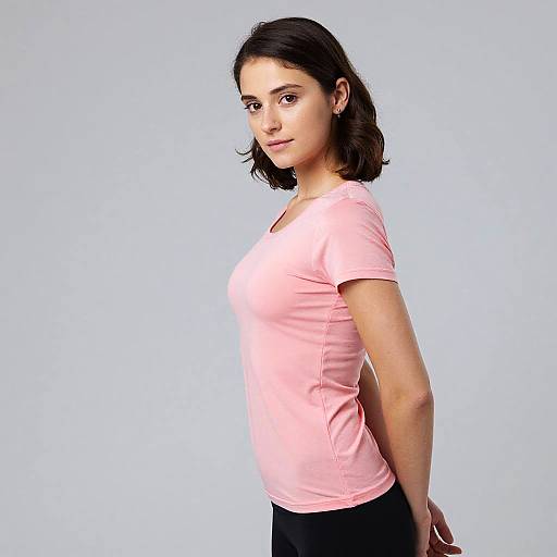 Photograph of a young woman with light skin and dark brown hair, wearing a pink t-shirt and black pants, standing in profile against a plain white