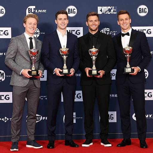 Photograph of four young men in suits, standing on red carpet, holding ABC awards, smiling, against black backdrop with ABC logos.