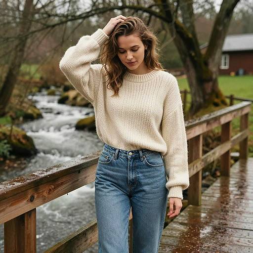 Photograph of a young woman with wavy brown hair, wearing a cream knit sweater and high-waisted blue jeans, standing on a wooden bridge