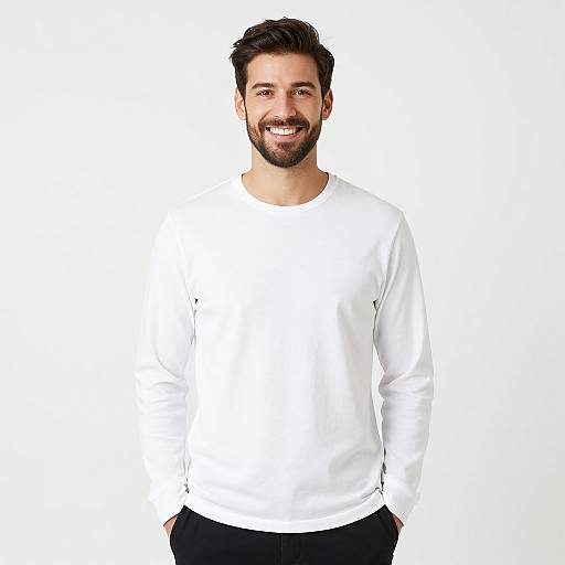 Photograph of a smiling, bearded man with dark hair, wearing a plain white long-sleeve shirt and black pants, standing against a white