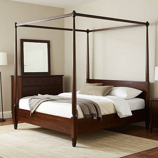 Photograph of a four-poster wooden bed with white linens, gray blanket, and pillows, set in a minimalist room with beige walls, dark